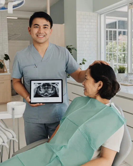 A smiling dentist holding a dental X-ray on a tablet beside a patient